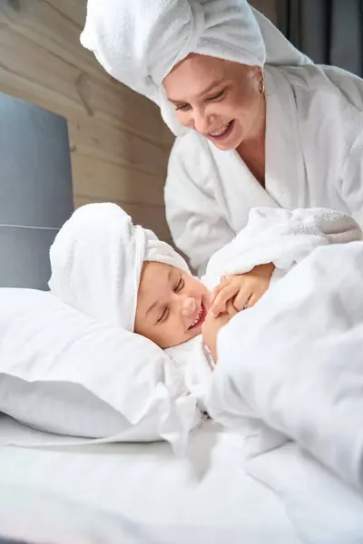 有魅力的妈妈穿着浴衣爱抚她漂亮的小女儿 她穿着柔软的浴衣躺在床上 头上披着大毛巾 女孩子们玩得很开心 — 图库照片