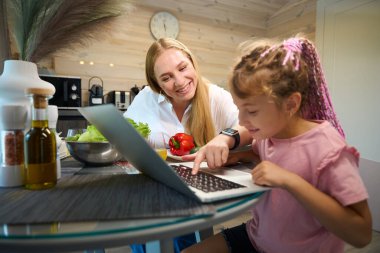 Güzel anne sebze salatası pişiriyor ve küçük kıza ev ödevini internet üzerinden yapmasına yardım ediyor. Aile akşamını birlikte geçiriyor.