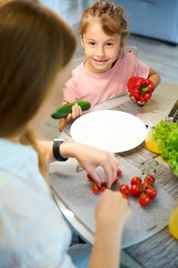 Küçük tatlı kız kameraya bakıyor ve annesi salatada domates keserken annesine kırmızı biber ve yeşil salatalık tutmasında yardım ediyor.