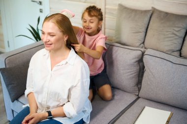Küçük gülümseyen kız saçlarını taramayı ve örmeyi öğreniyor, güzel sarışın annesine, kızına ve annesine saç stili yapıyor.