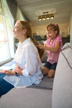 Annesi samimi bir şekilde gülerken, küçük kızı koltukta oturup annesinin saçını örmeye çalışıyor, kızlar kuaförde oynuyor, hobi yapıyor.