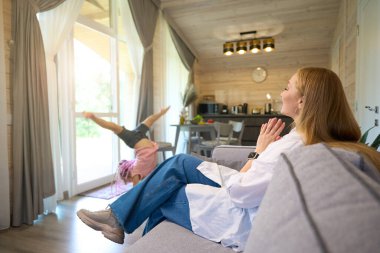 Anne kanepede oturuyor ve kızına bakıyor. Performans gösteriyor, takla atıyor, kafa üstünde duruyor, atletizm yapıyor, anne içtenlikle alkışlıyor.