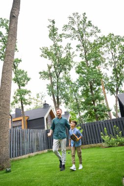 Baba ve oğul evin yakınındaki yeşil çimlerin üzerinde yürüyorlar. Beyzbol aletleri var.