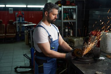 İş kıyafetleri içinde ciddi bir araç tamircisi atölyedeki yedek kulübesinde metal araba parçalarını öğütüyor.