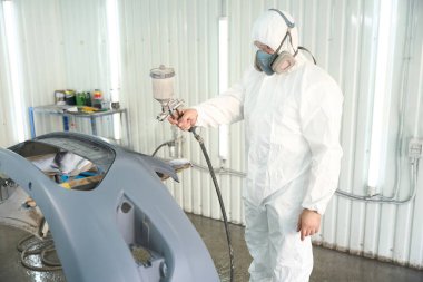Korumalı giyimli otomotiv tamircisi atölyedeki müşteri aracı tamponlarını boyamak için sprey tabanca kullanıyor.