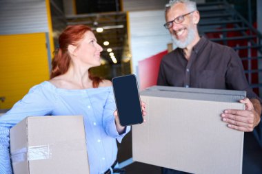 Kadının elinde cep telefonu var. Kocasıyla birlikte bir depodalar.