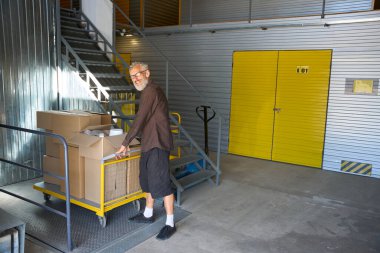 Sakallı adam kargo arabasının yanında duruyor. Arabada bir sürü karton kutu var.