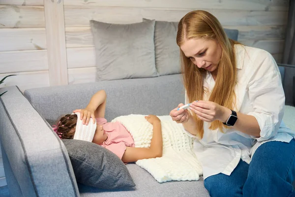 金发母亲困惑于她躺在沙发上的小女儿的体温过高 额头被压得很紧 有感染考拉病毒的危险 — 图库照片