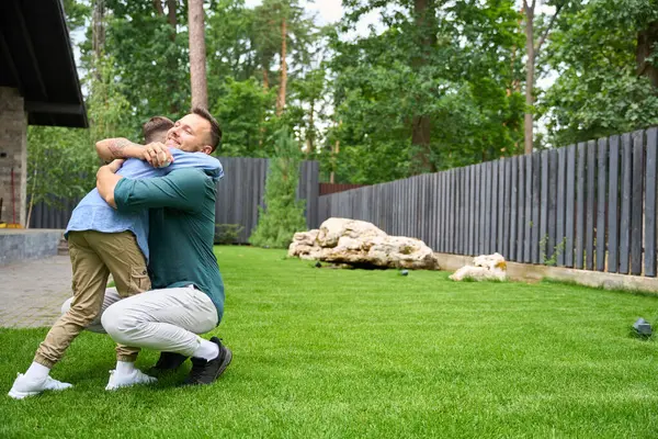 父亲笑着把儿子抱在房子的草坪上 男孩戴着棒球手套 — 图库照片
