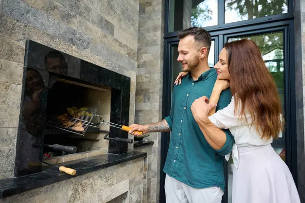 Unshaven man is cooking food over a fire on the terrace, next to his beautiful wife