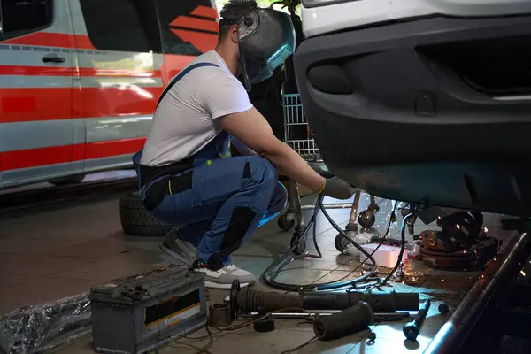 Automotive technician in protective helmet and gloves welding car body parts in garage