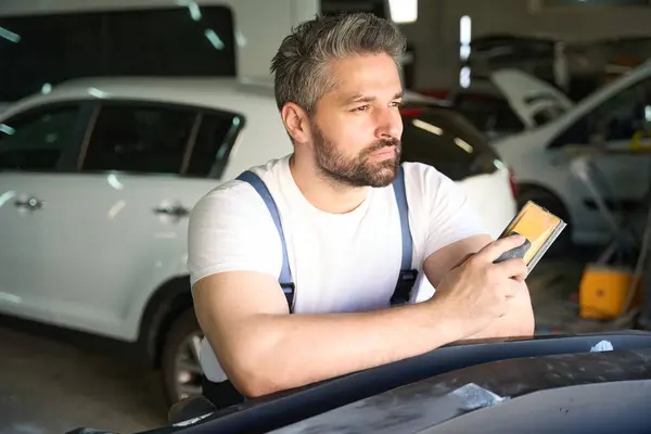 Thoughtful car repair shop worker leaning on vehicle bumper cover with sanding block in hands