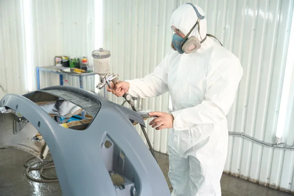 Automotive repair shop worker dressed in protective clothing painting automobile bumper with spray gun