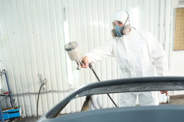 Serious refinish technician in protective clothing applying paint to automobile bumper with spray gun