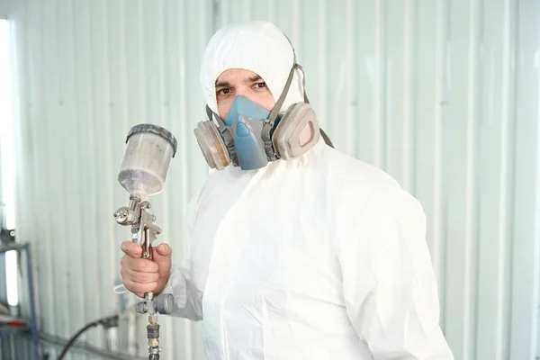 Portrait of paint technician in protective clothing and with respirator holding spray gun in his hand