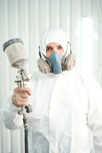 Waist-up portrait of painter with respirator and in protective clothing holding spray gun