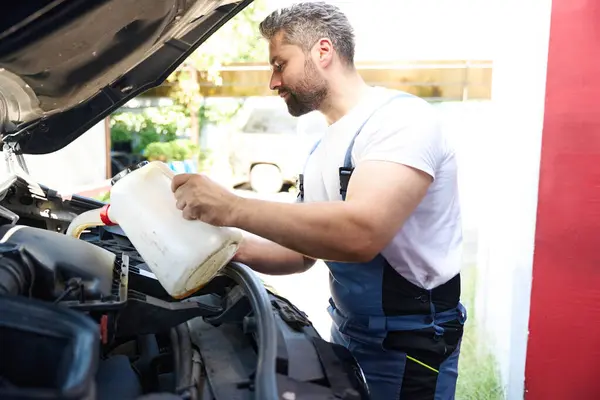 Focused mechanic pouring motor oil from plastic tank into automotive engine while standing at open car bonnet