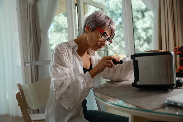高齢女性は台所のテーブルでトースターを修理しています 彼女はスクリュードライバーを使用しています — ストック写真