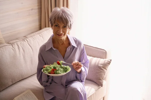 本と軽い食べ物 ライラックの服の女性と居心地の良いソファーに座っているかわいい老婦人 — ストック写真