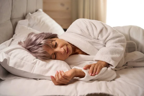 Elderly woman sleeps sweetly in a bright bedroom on soft pillows, a woman in a soft bathrobe