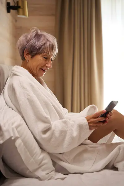 Happy pensioner in a bathrobe sits in bed with a mobile phone, she sits in soft pillows
