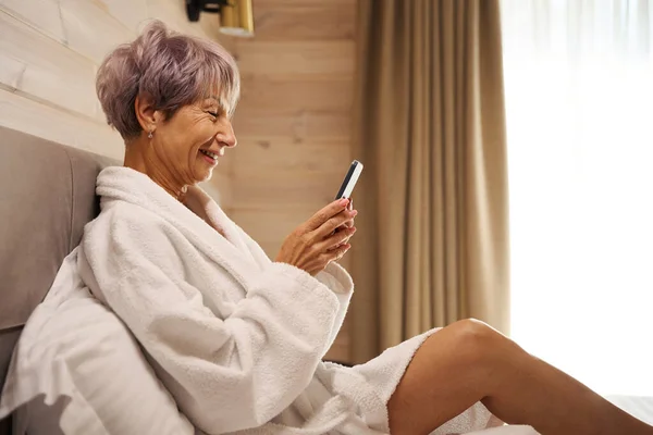 精力充沛的老太太在卧室里用手机交流 坐在柔软的枕头上 — 图库照片