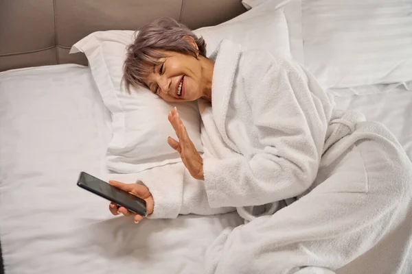 Smiling elderly woman resting in bed with mobile phone, she lies on fluffy pillows