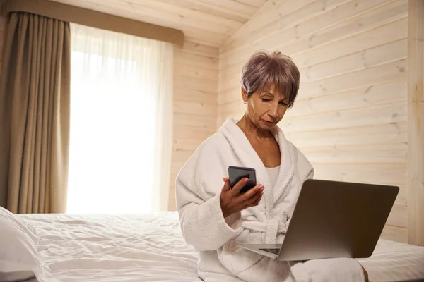 Pensionerは携帯電話で通信し ラップトップを使用し ベッドルームのベッドに座っている — ストック写真