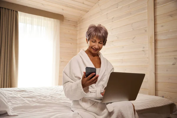Nice lady communicates by mobile and uses a laptop, she sits on the bed in the bedroom