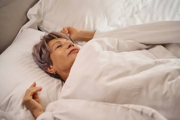 Gray-haired woman is dozing in a soft bed, she lies on fluffy pillows