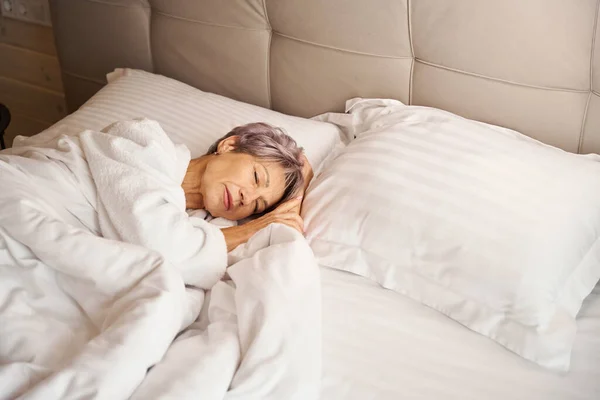Gray-haired woman is dozing in a large bed, she lies on fluffy pillows