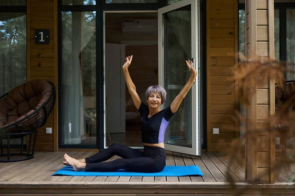 Joyful lady performs physical exercises on a wooden terrace, she sits on a roll mat