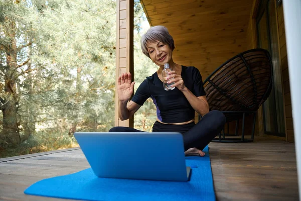 Elderly woman in sportswear communicates online, she sits on a roll mat on the terrace