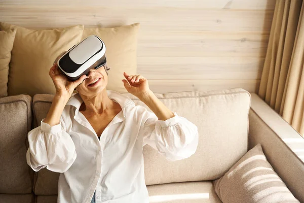 女性は仮想ゲームをプレイする部屋のソファーに座って 彼女は仮想現実のメガネを使用しています — ストック写真