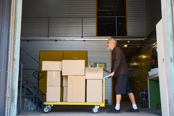 Bearded male is pushing a cargo cart around a warehouse, there are a lot of cardboard boxes on the cart
