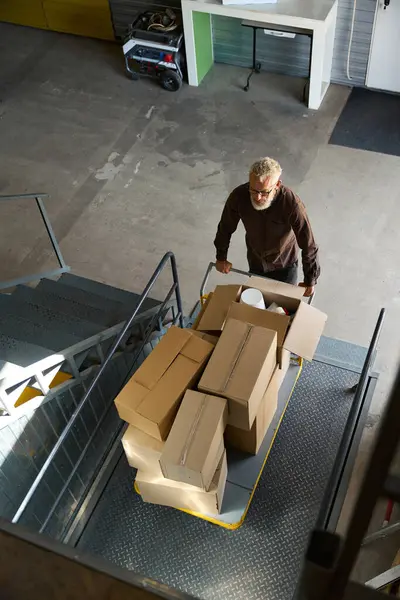 Gray-haired male lifts cart with things on a freight elevator, there are a lot of cardboard boxes on the cart