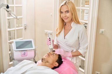 Kliniğin genç müşterisi RF yüz gerdirme prosedürüne tabi tutuluyor, doktor alın bölgesinde çalışıyor.