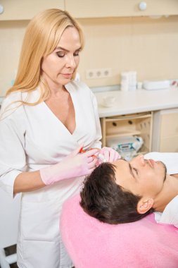 Kozmetik kliniğinde güzellik iğnesi yaptıran genç adam, hastalarla ilgilenen doktor