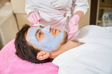 Kahverengi saçlı bir adam kozmetoloji ofisinde rahat bir kanepede yatıyor. Kozmetik uzmanı yüzüne temizleyici bir maske takıyor.