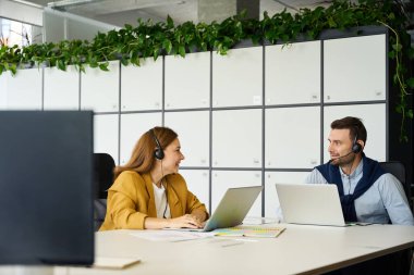 Bir erkek ve bir kadın büyük bir ofis masasında oturuyorlar, insanlar dizüstü bilgisayar ve kulaklık kullanıyorlar.
