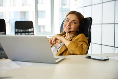 İş yerinde masa başında çalışan genç bir kadın, bir dizüstü bilgisayar ve kulaklık kullanıyor.