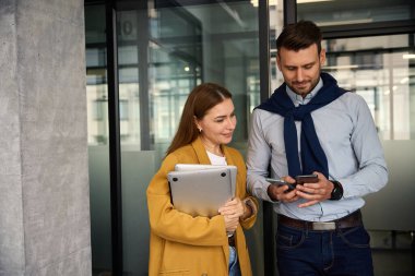 İş arkadaşları ofis koridorunda iletişim kuruyor, cep telefonları, dizüstü bilgisayarları ve belgeleri var.
