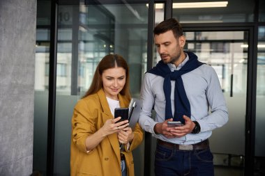 Ofis koridorunda cep telefonları olan bir adam ve bir kadın, oda temiz ve aydınlık.