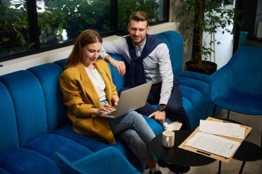 Meslektaşlar ortak çalışma alanındaki belgelerle çalışırlar. Yumuşak mavi bir koltukta, bir kadının dizüstü bilgisayarı vardır.