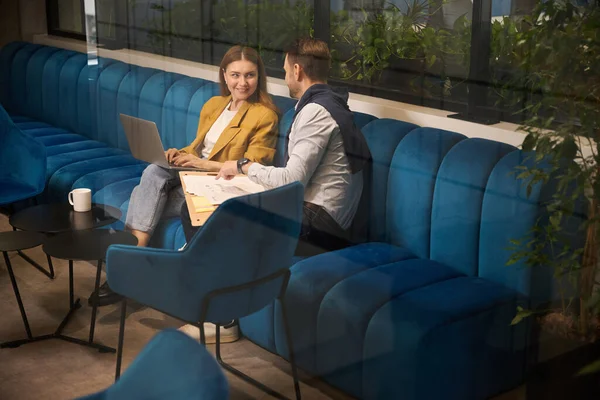 女性和男性在一个软软的蓝色沙发上 穿着黄色夹克的女性工作时 在一个共同工作的空间里处理文档 — 图库照片