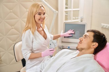 Kadın kozmetikçi, müşteriyi smas kaldırma prosedürü için cihazla tanıştırır, müşteri kozmetoloji kürsüsünde oturur.