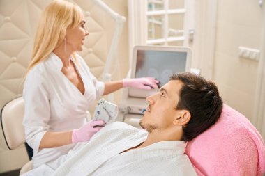 Kadın, müşterisine smas kaldırma işlemi için bir cihaz sunar, erkek ise kozmetik kürsüsünde oturur.