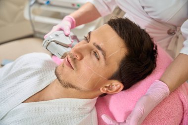 Uzman yanak bölgesindeki bir müşteriye kozmetik donanım prosedürü uyguluyor. Adam kozmetoloji kürsüsünde oturuyor.