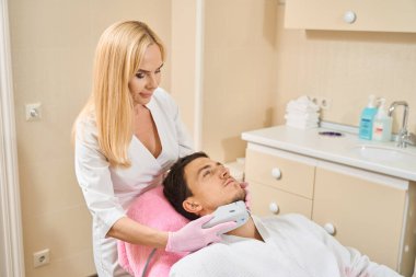 Doktor çene bölgesindeki bir müşteriye mikronedle RF uyguluyor. Adam kozmetik kürsüsünde oturuyor.