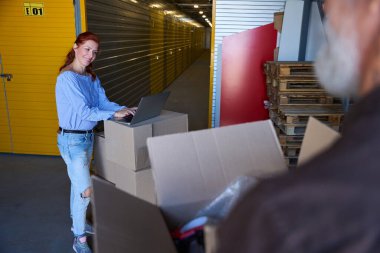 Depoda dizüstü bilgisayar kullanan hoş bir kadın. Yanında kutuları olan gri saçlı bir adam var.